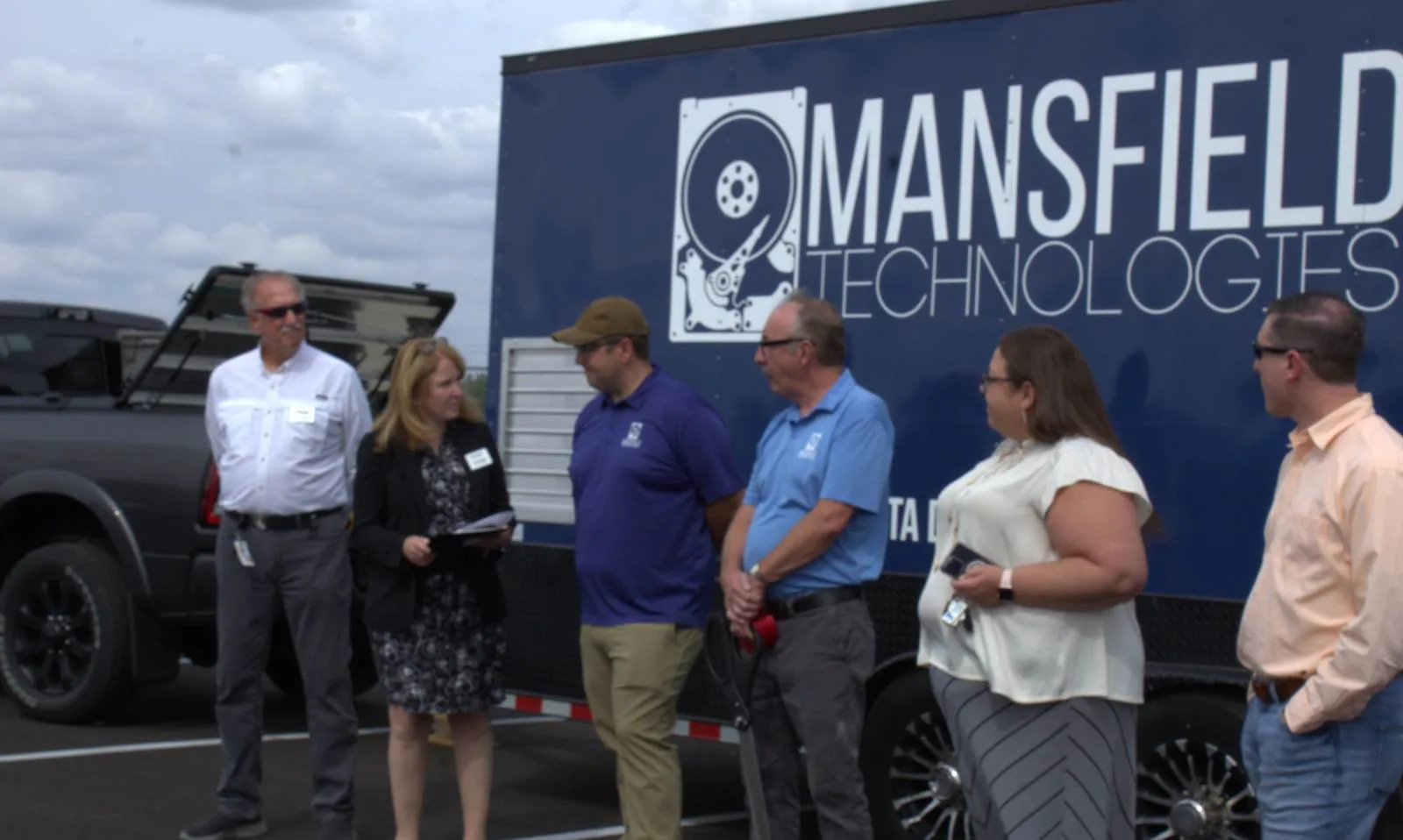 Six people stand in front of a navy blue Mansfield Technologies mobile shredding trailer at a ribbon cutting